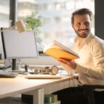 A man smiling and holding a book while sitting on his office chair.