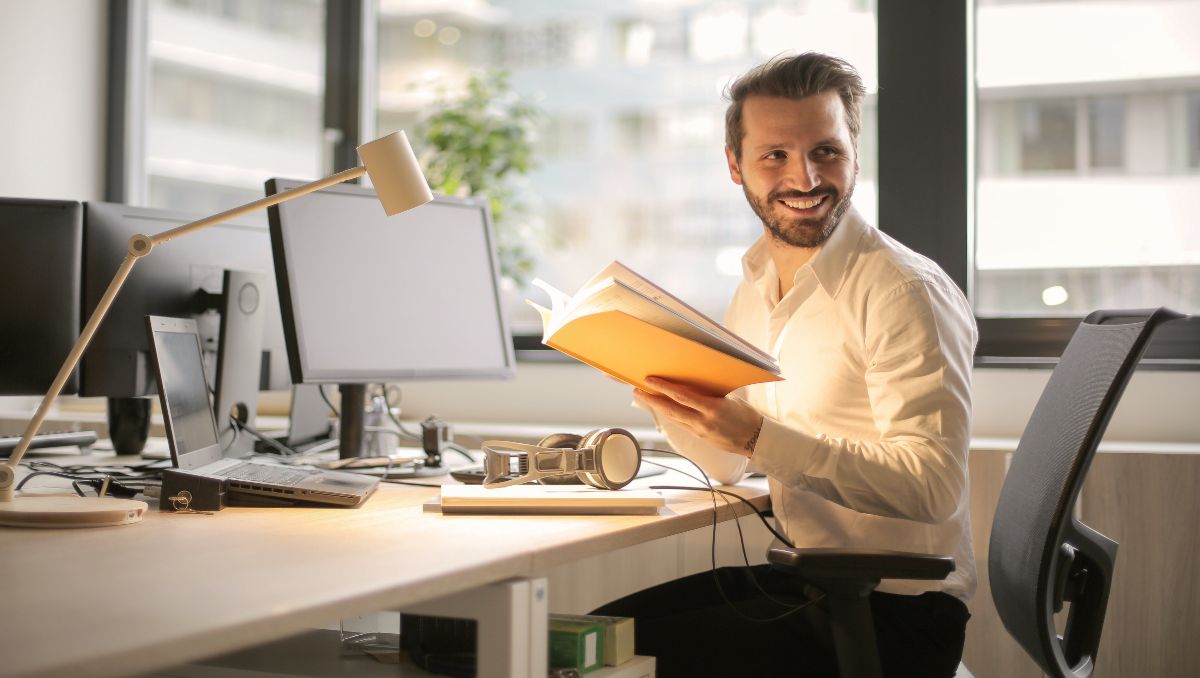 A man smiling and holding a book while sitting on his office chair.