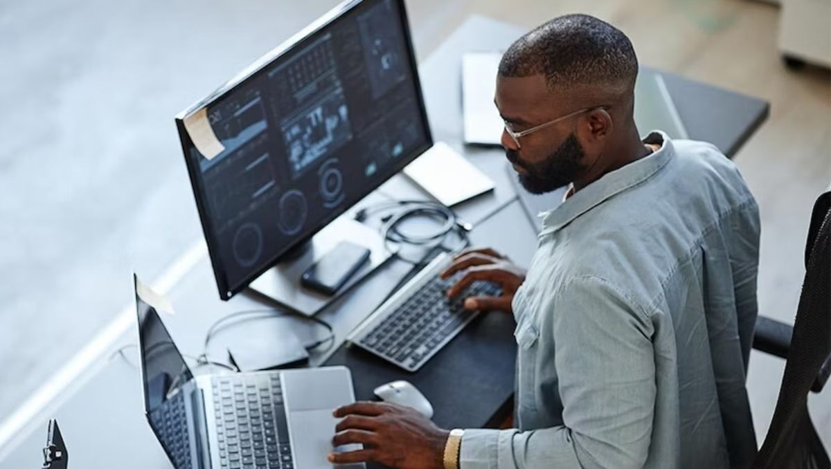 A male office worker sitting on his chair while typing on a laptop and desktop computer. 