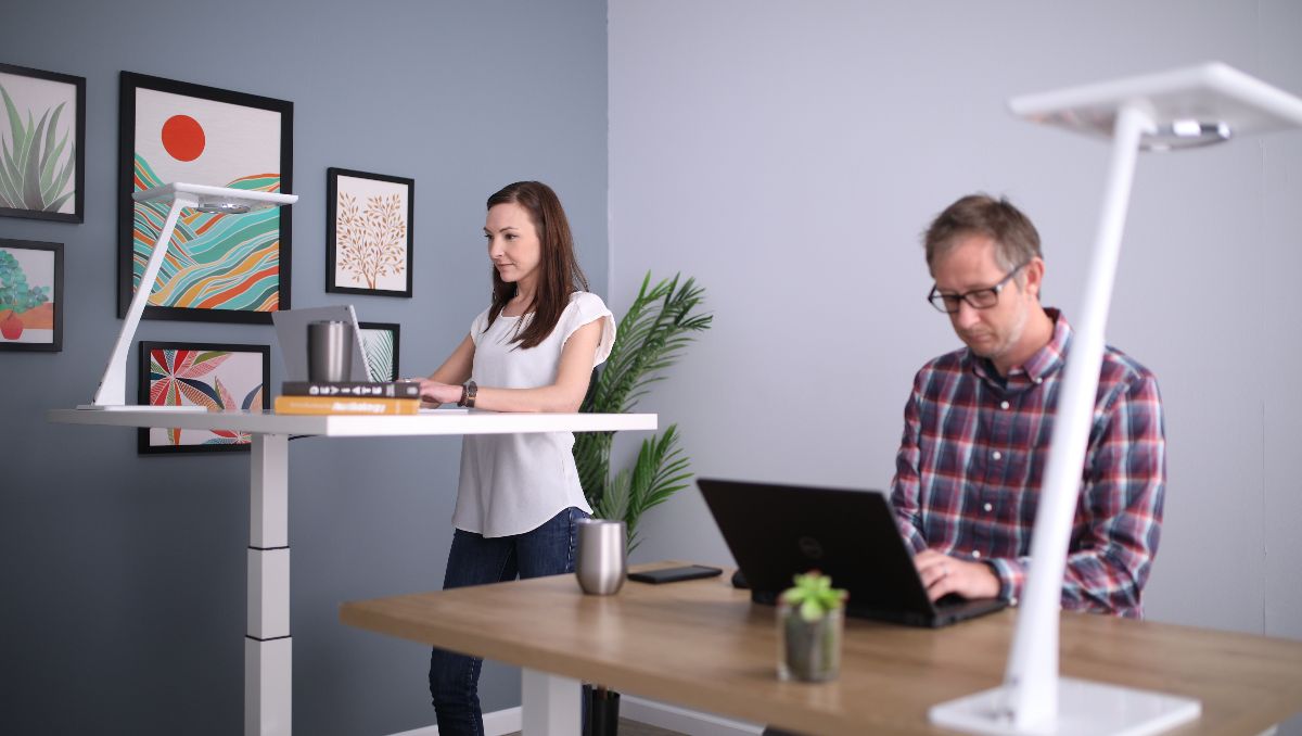 A woman working at a standing desk and a man sitting on his desk while working.
