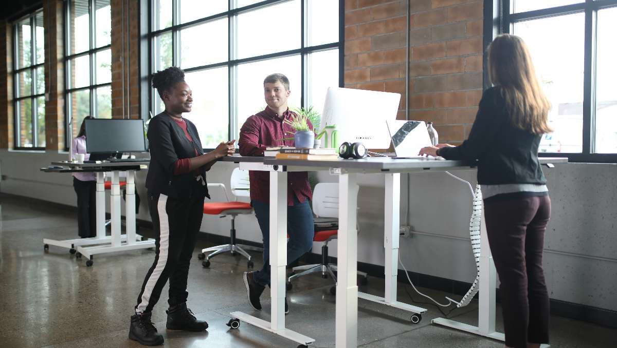 Two women and one man talking while working at their standing desks.