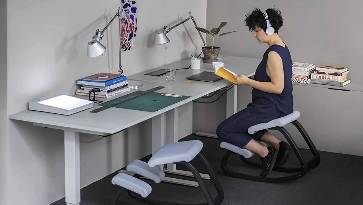 A woman reading a book and sitting on a kneeling chair.