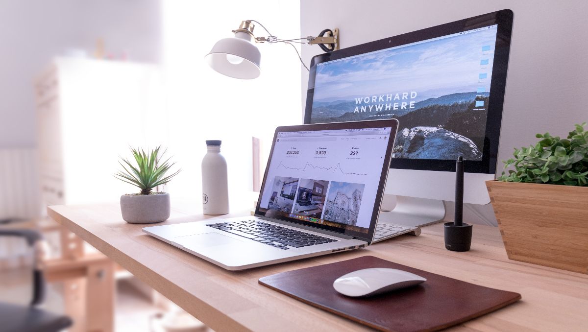 A wooden desk with laptop, desktop, mouse, plant, and a water bottle on top.