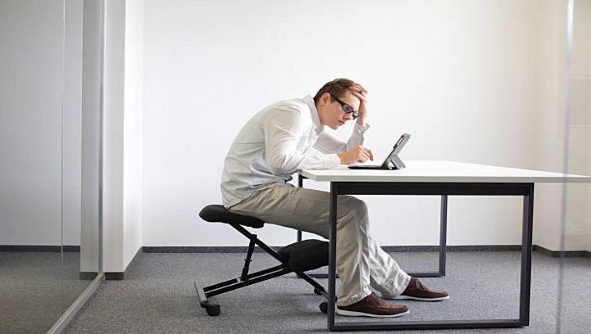 A man slouching on the desk while sitting on a kneeling chair.