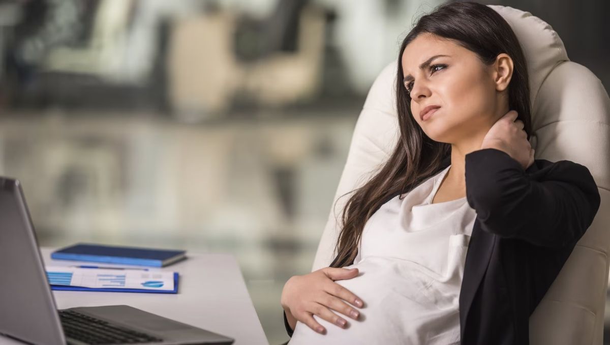 A tired pregnant woman sitting in an office chair while holding the side of her neck.