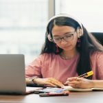 Student writing on a desk facing laptop and wearing headphones