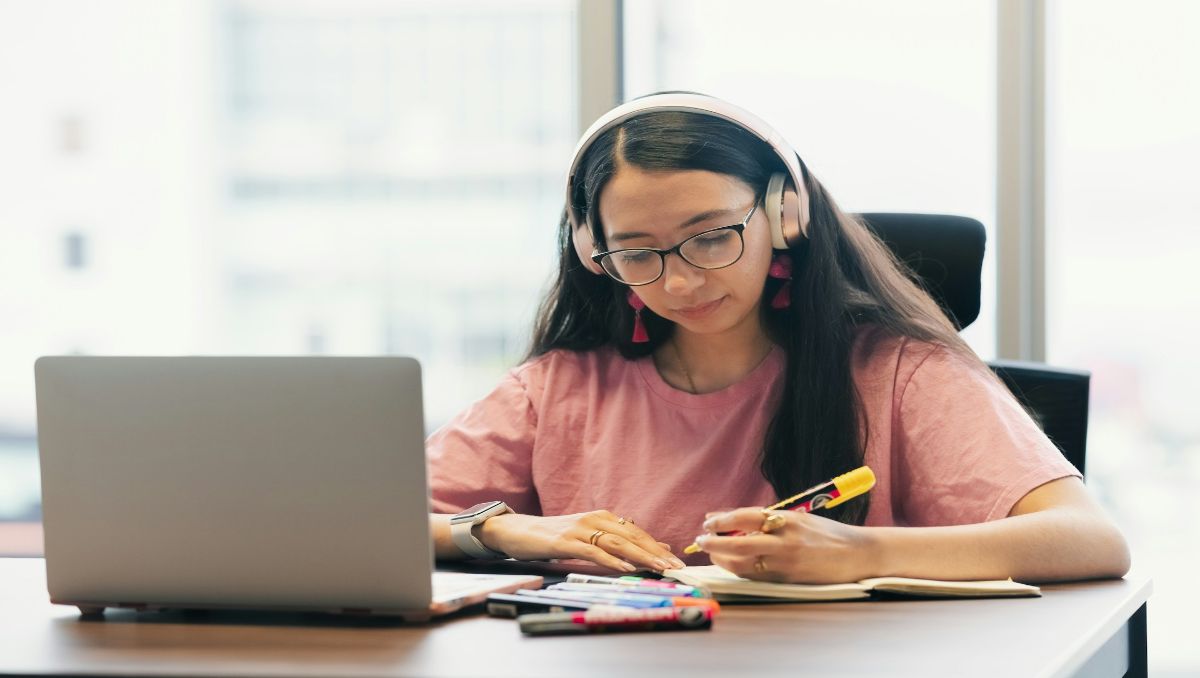 Student writing on a desk facing laptop and wearing headphones